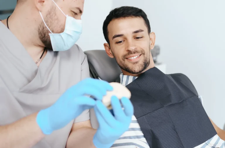 Masked dentist in blue gloves holds a dental model beside a smiling man reclining in a dental chair