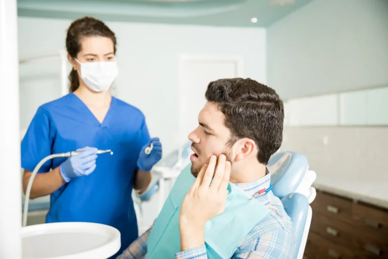 Masked dental professional in blue scrubs examines a male patient holding his jaw in a dental office