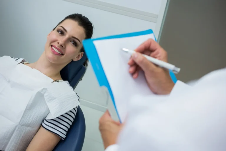 Smiling dark-haired woman in a striped top at a dental office as a clinician writes notes on a clipboard
