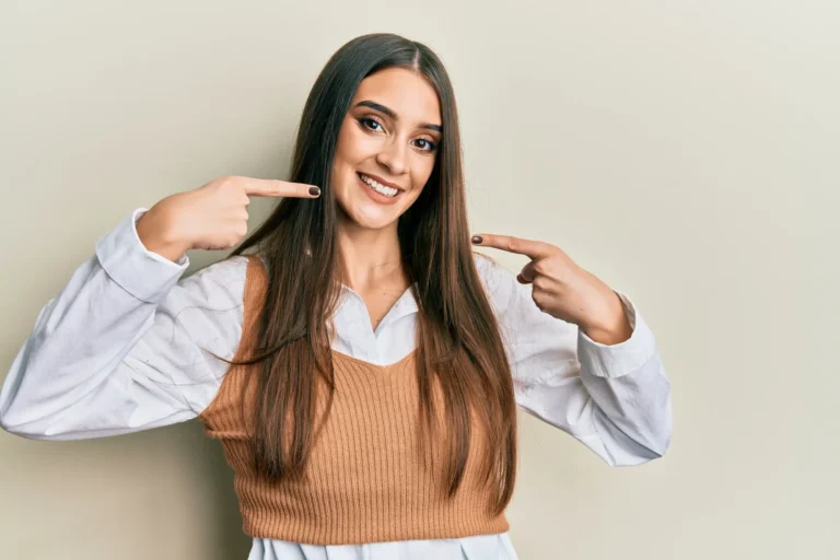 Woman with long brown hair in a knit sweater smiling and pointing at her teeth with both index fingers